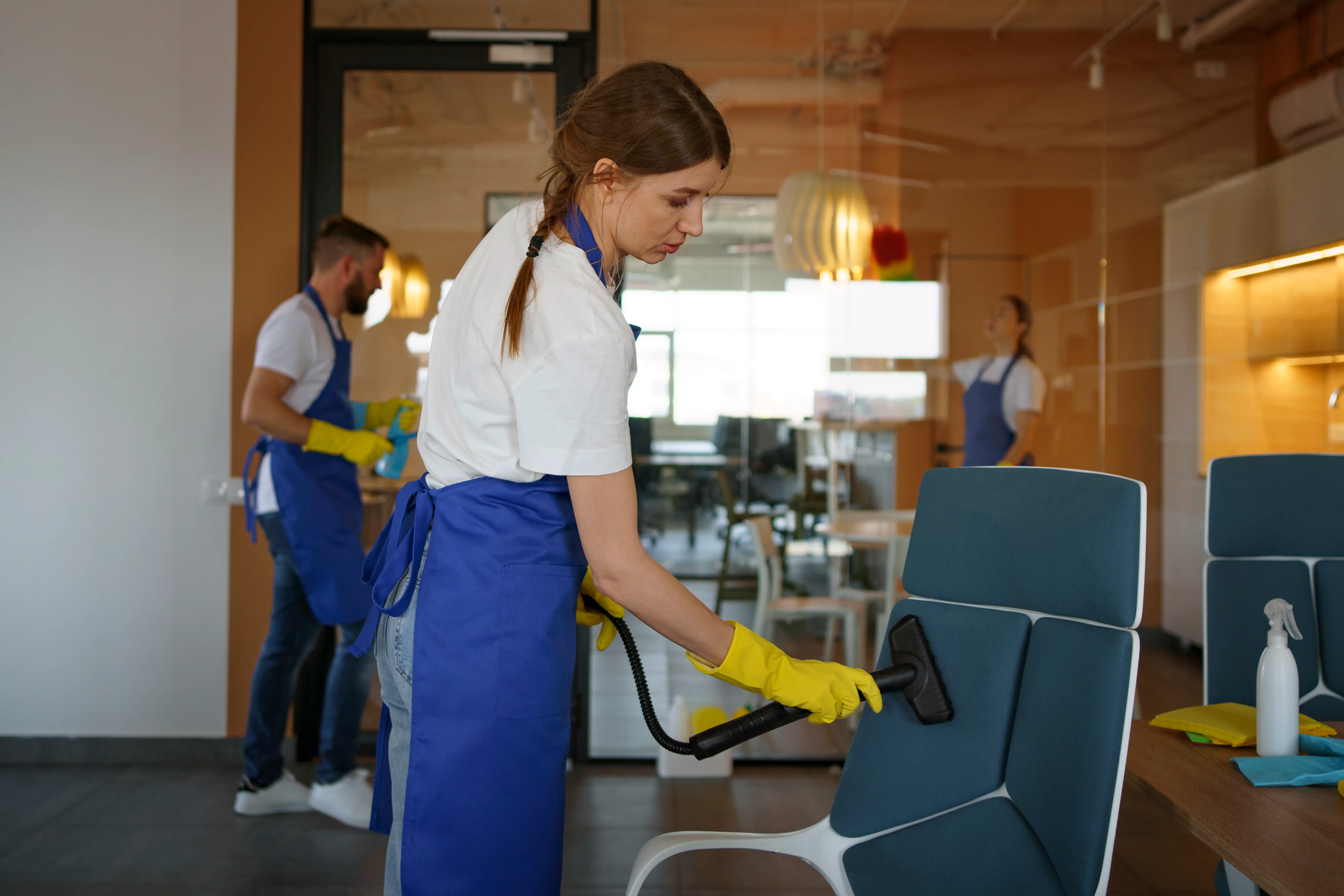 A woman wearing a blue apron and rubber gloves is cleaning a chair with a cloth