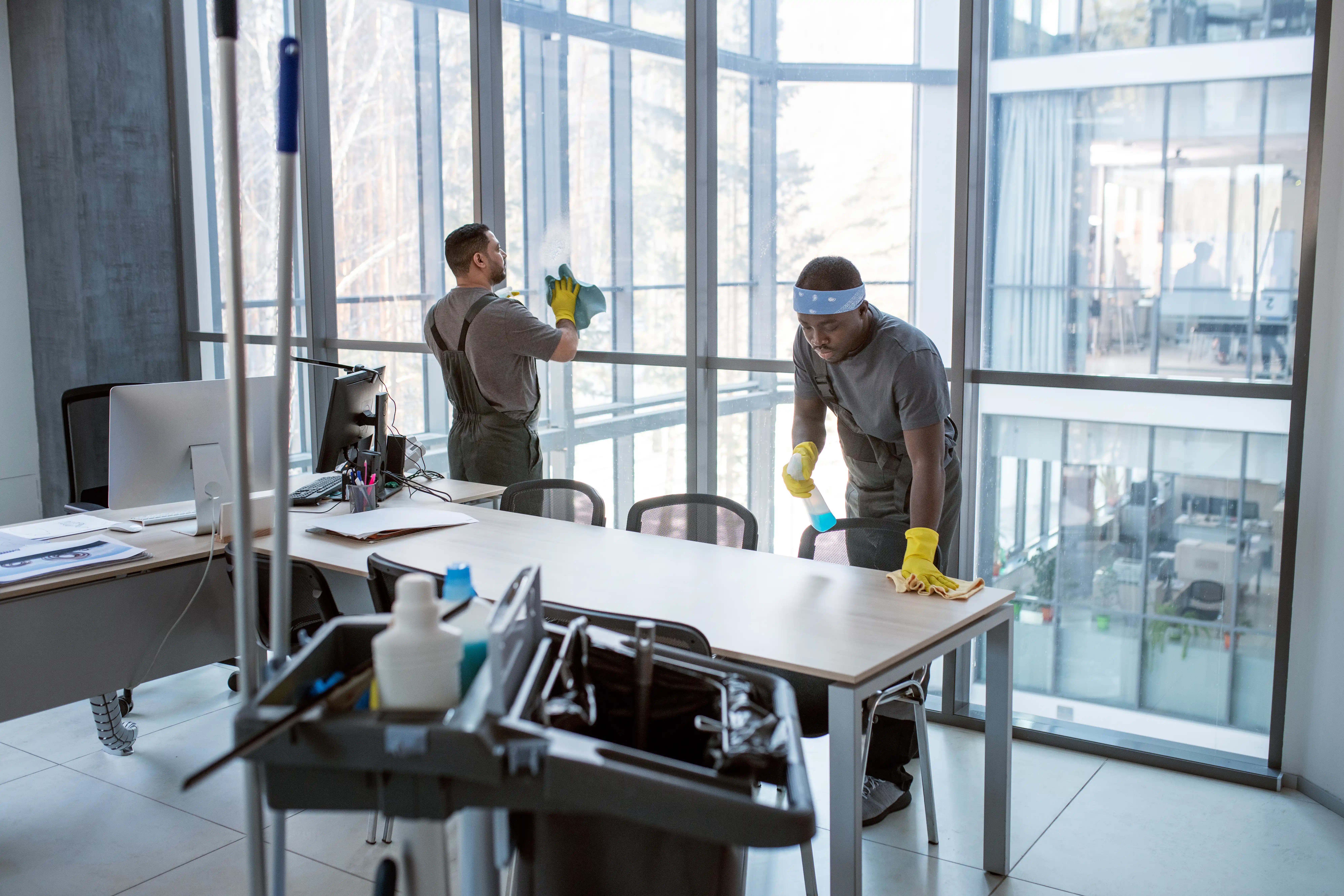 Two workers in gray uniforms and yellow gloves clean a modern office. One wipes large windows, the other cleans a desk. A cleaning cart is in the foreground.